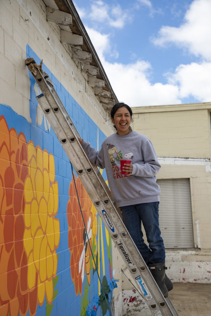 Joleen Jewell stands on a ladder holding a cup of paint. The ladder leans against a wall that she has painted with red and yellow flowers against a blue sky.