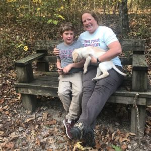 A woman sits on a bench in the woods. She has her arm around a kid, and a yellow puppy in her lap.