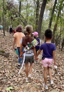 A group of girls walk on a trail through woods. One girl wears a vest with several girl scout badges