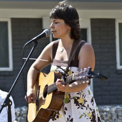 A woman with dark hair plays a guitar and sings at a microphone