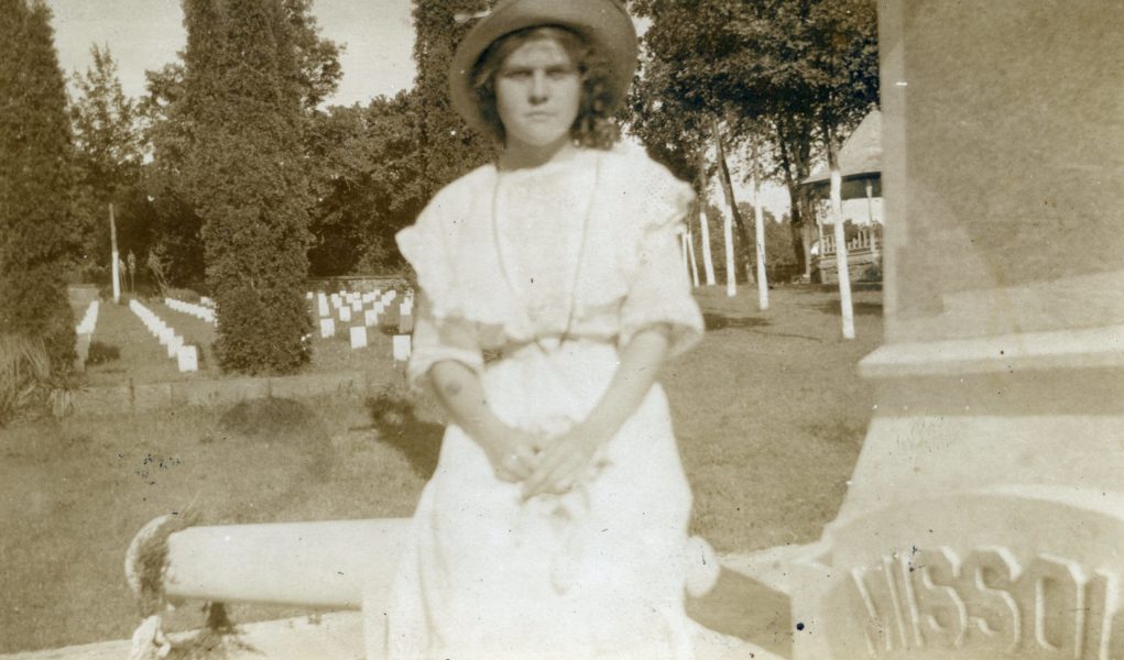 Belle Dinwiddie, a young white woman in a white dress wearing a straw hat sits on cement canon at Confederate Cemetery in Fayetteville, circa 1915. Her expression is neutral and she looks directly at the camera.