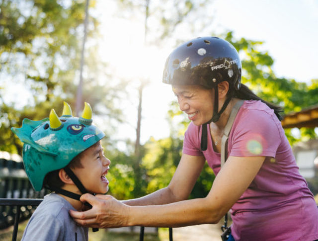 Noy, a Lao woman, helps her young son Ollie to put on a bicycle helmet. She is grinning and he is laughing.