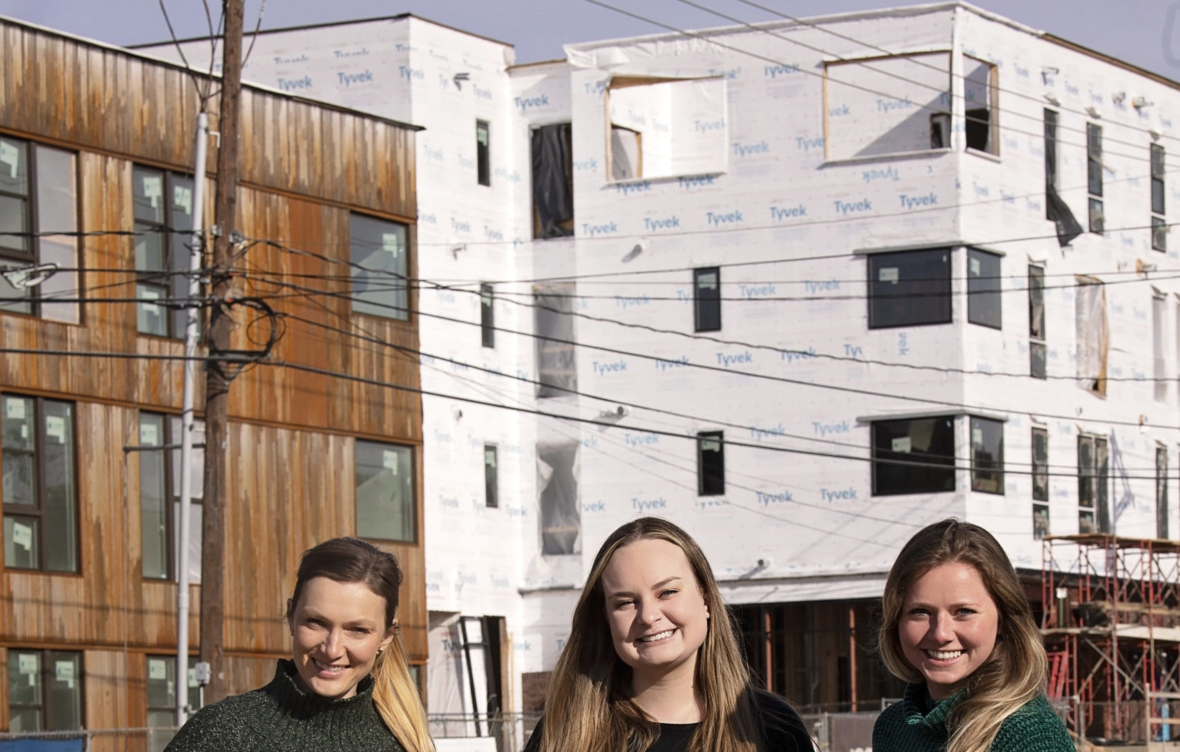 Construction manager Kaitlyn Fondano, Operations Manger Paige Sauerwein, and Project Architect Kiara Luers stand in front of an under construction apartment building