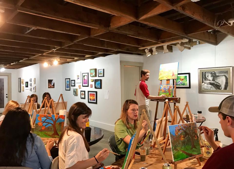 Alex Cogbill stands at an easel and paints a colorful landscape painting. In the foreground people each paint at easels.