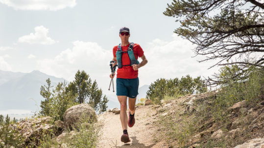 A man runs toward us on a trail on a mountainside. He is wearing a red shirt and shorts and a backpack.