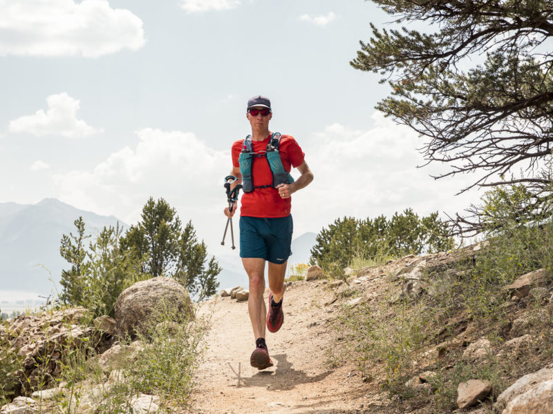 A man runs toward us on a trail on a mountainside. He is wearing a red shirt and shorts and a backpack.