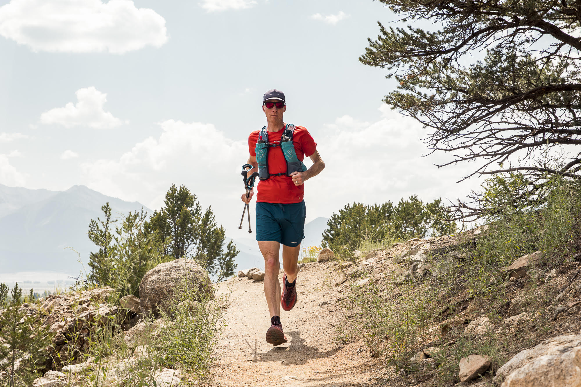 A man runs toward us on a trail on a mountainside. He is wearing a red shirt and shorts and a backpack.