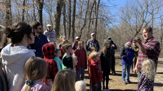 Outdoors under bare winter trees, a man in a flannel shirt holds out a turtle in an open hand to a gathering of kids and parents