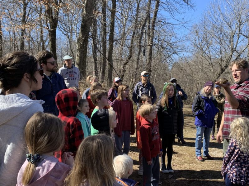 Outdoors under bare winter trees, a man in a flannel shirt holds out a turtle in an open hand to a gathering of kids and parents