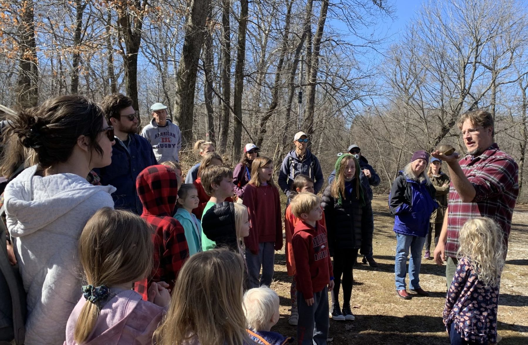 Outdoors under bare winter trees, a man in a flannel shirt holds out a turtle in an open hand to a gathering of kids and parents
